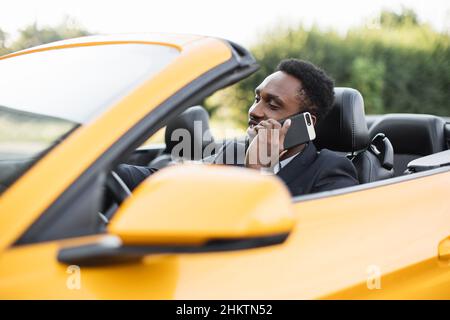 Handsome African businessman talking on the mobile phone while sitting on back seat of a sport yellow car. African American male business executive travelling by a car and making phone call. Stock Photo