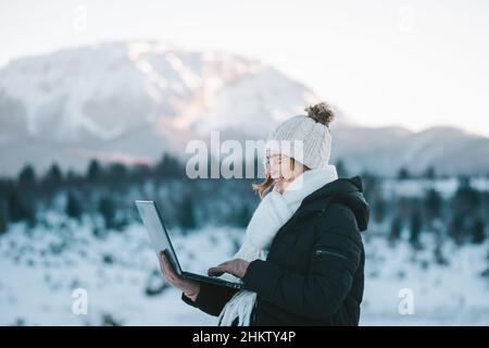 Smiling Caucasian female freelancer using laptop in snowy outdoors with mountain on background Stock Photo