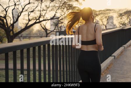 Woman in sportswear wearing a protective mask jogging exercise on the bridge in the city at sunset. Stock Photo