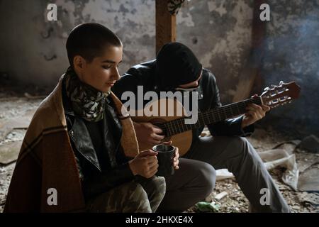 a young adult woman and a man together in a destroyed building. The guy in the mask plays the guitar, the girl is wrapped in a blanket. The concept of survival in a post-apocalyptic world. Stock Photo