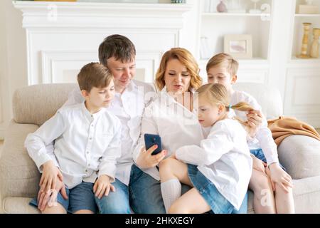 Happy parents with three kids using smartphone at home together sitting on cozy sofa in living room, smiling mother and father with two sons and daughter looking at phone screen taking selfie cheerful Stock Photo
