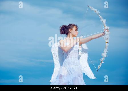 Teen girl in white angels dress, angel cupid valentin shoot with bow arrow. Teenager with feathers wings, Valentines Day. Stock Photo