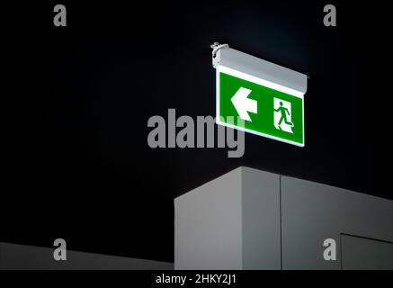 Fire exit sign. Green electric emergency evacuation escape sign with illuminated light on the black ceiling in the dark building with copy space. Stock Photo