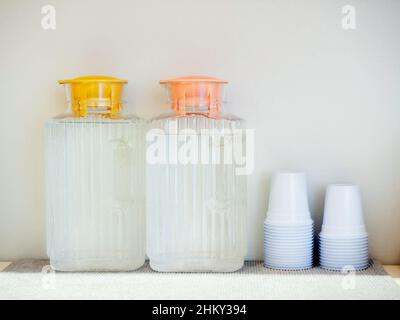 Two plastic bottles with yellow and pink pastel lids full of cold water were placed near the stack of white plastic cups on the shelf to serve custome Stock Photo
