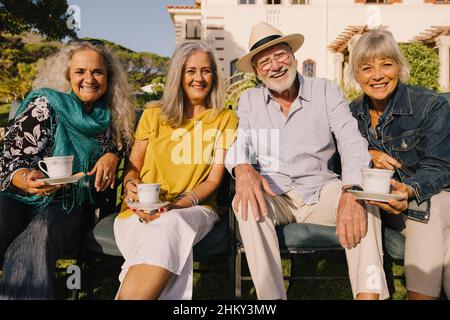 Happy senior citizens smiling at the camera while relaxing outside a retirement home. Group of carefree elderly people having tea together and enjoyin Stock Photo