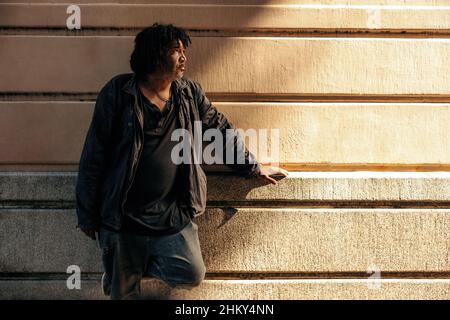 Homeless man looking away thoughtfully while standing alone outdoors during the day. Middle aged unemployed man having a reflective moment while stand Stock Photo