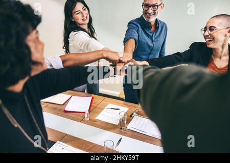 Ecstatic group of businesspeople bringing their fists together while standing in a boardroom. Diverse group of businesspeople smiling cheerfully while Stock Photo