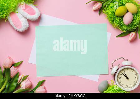 Easter composition. Eggs in a nest tulips flowers bunny ears alarm clock on a pink background. Empty sheet of paper with space for text and copy space Stock Photo