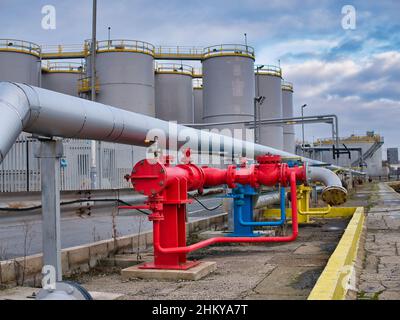Brightly coloured industrial pipelines,  control valves and a low yellow wall with a background of large, tall grey storage tanks. Stock Photo