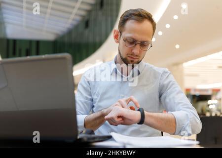 Using smart fitness bracelet at work. Businessman sitting at table and touches smart watch. Notification on gadget Stock Photo