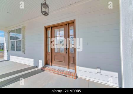 Porch of a white house with wooden siding and windows. There is a ceiling lamp at the front of the wooden door with two side glass panels near the win Stock Photo