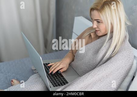 Sick ill young woman feel cold covered with blanket sit on sofa watching movie on laptop, annoyed girl shiver freezing warming at home wrapped with Stock Photo