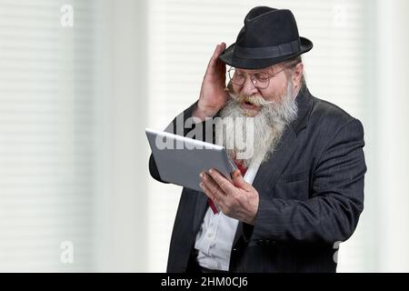 Old senior businessman looking at digital tablet pc with surprise expression. Shocked bearded man using computer tablet standing indoor. Stock Photo