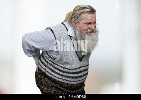 Old man with gray beard suffering from backache on blurred background. Unhealthy senior man close up. Stock Photo