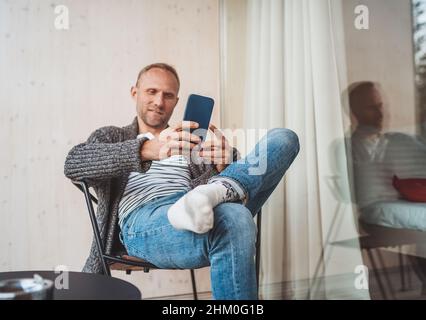 Smiling middle-aged man dressed open cardigan, jeans, and warm socks sitting on house balcony and smiling when he using modern smartphone. Everyday li Stock Photo