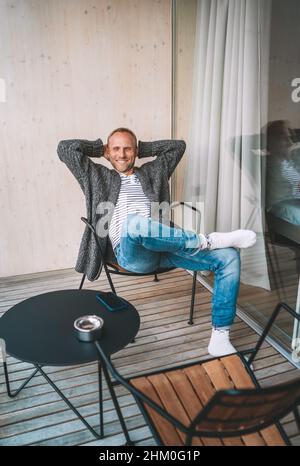 Portrait of a Middle-aged man dressed open cardigan, jeans, and warm socks sitting on forest house balcony and enjoying the fresh air. Everyday lifest Stock Photo