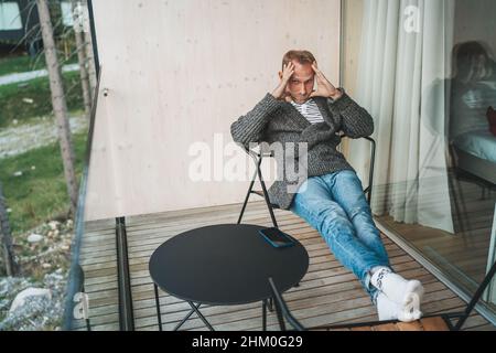 Portrait of a tired Middle-aged man dressed in an open cardigan, jeans, and warm socks sitting on forest house balcony. He massaging temples to increa Stock Photo