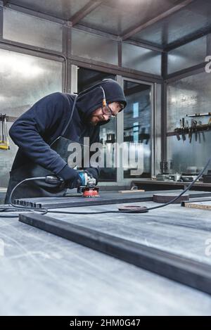 Worker, wearing special protective gloves and glasses, using grinder to polish piece of metal frame pressed to work table. Man holding hand grinder. Vertical shot Stock Photo