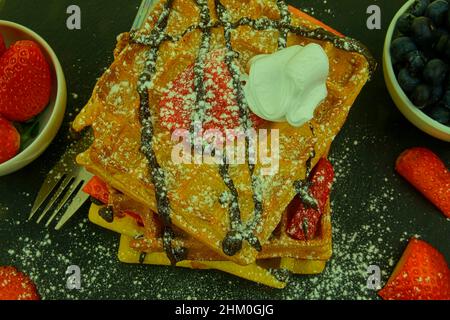 Square Belgian waffles with berries and icing sugar on a black plate. Tasty sweet sugary waffles. Melted chocolate and whipped cream on waffles. Top Stock Photo