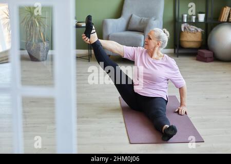 Senior active woman in t-shirt and leggins sitting on mat and doing stretching exercise for legs in living-room Stock Photo