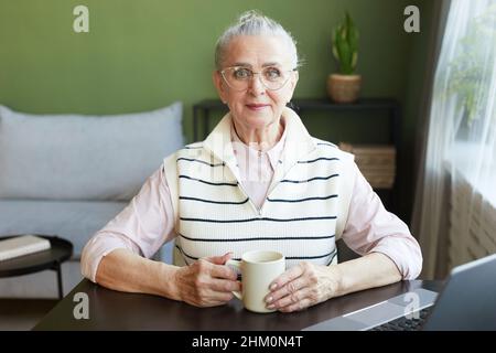 Happy mature female in eyeglasses and casual clothes having tea while sitting by table in front of laptop in living-room Stock Photo