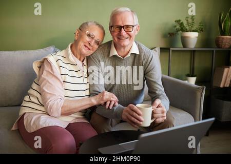 Happy and affectionate senior husband and wife sitting on sofa in front of laptop in living-room and looking at camera Stock Photo