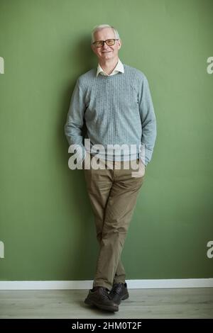 Happy elderly man in eyeglasses, casual brown pants and light blue pullover standing by green wall and looking at camera Stock Photo