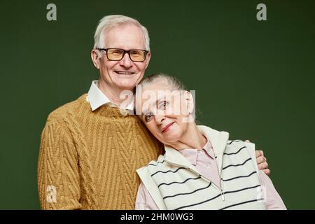 Happy elderly husband and wife in pullovers standing close to one another against dark green background in front of camera Stock Photo