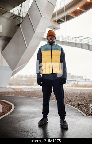 Young serious sportsman of African ethnicity wearing warm tracksuit and sleeveless jacket standing on asphalt road outdoors Stock Photo