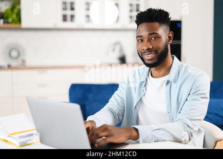 Portrait of dark-haired man using laptop in home office. Brunette male entrepreneur looking at the camera and smiling toothy, kitchen on the background Stock Photo