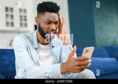 Upset multiracial businessman is talking on the smartphone. Portrait of sad failed serious male employee having conversation via video call while sitting at the sofa. Unsuccessful negotiation concept Stock Photo