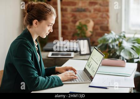 Young confident businesswoman in formal clothes typing on laptop keyboard while analyzing financial data on screen Stock Photo