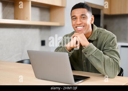Confident concentrated latin freelancer guy working on laptop at home office, while sitting at desk and using computer. Multiracial eastern man looking at the camera and enjoying remote work Stock Photo