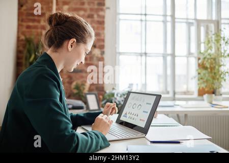 Young contemporary female accountant looking at financial data on laptop screen while analyzing it by desk in openspace office Stock Photo