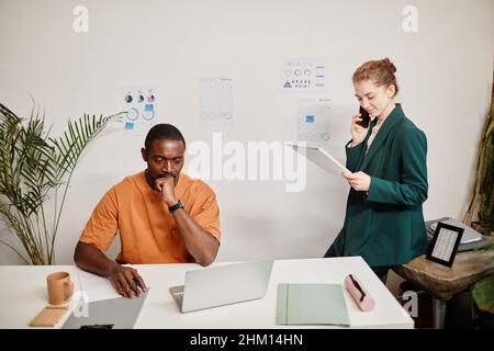 Young serious African American businessman looking at laptop screen while his Caucasian female colleague consulting client on mobile phone Stock Photo