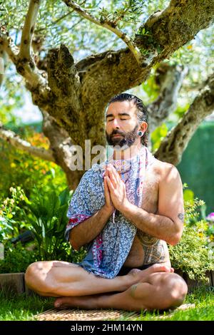 Young bearded man unclothed practicing yoga (lotus pose) outside in a garden close to an olive tree. Stock Photo