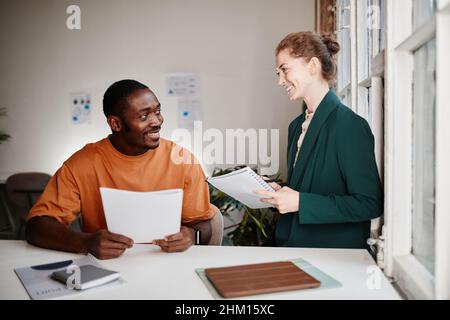 Two happy young intercultural economists planning work and discussing financial documents at working meeting in bureau Stock Photo
