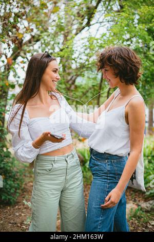 Two young teenager girls listening to music on white headphones and having fun at a park. Stock Photo