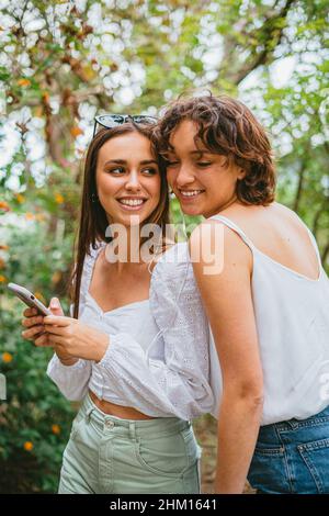 Two young teenager girls listening to music on white headphones and having fun at a park. They are smiling at each other. Stock Photo