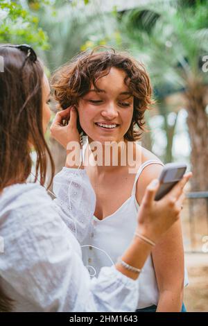 A teenager girl is helping her friend to put an ear-bud. They are having fun at the park. Stock Photo