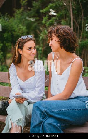 Two smiling teenage girls listening to music while sitting on a wood bench at the park. They are looking at each other. Stock Photo