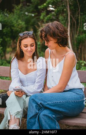 Two teenage friends looking at the phone and listening to music while sitting on a bench at the park. Stock Photo