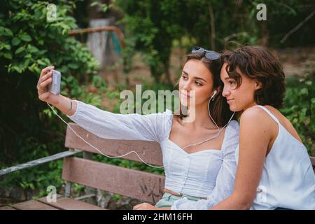 Two teenage girls taking a selfie while sitting on a bench at a park. Stock Photo