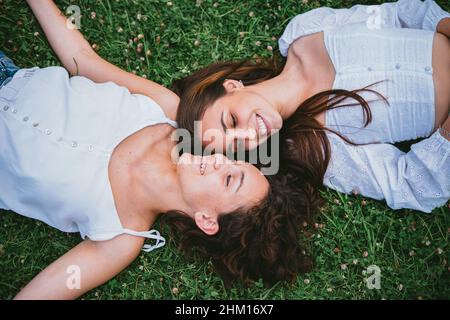 Two teenager girls smiling and looking at each other while they are laying down on grass in a park. They seem relaxed. Stock Photo