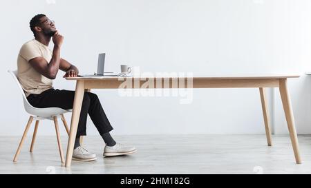 Pensive young black guy using laptop, sitting at desk, thinking over something, working online from home office Stock Photo