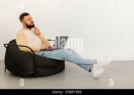Thoughtful Man Using Laptop Thinking Sitting In Chair At Home Stock Photo