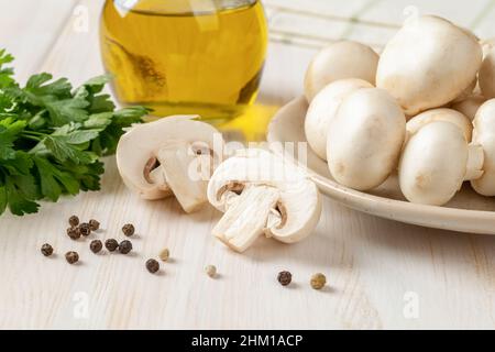 Close-up of raw champignon cut in half, whole button mushrooms, green parsley and olive oil jar on a white wooden table. Cooking fresh white mushrooms Stock Photo