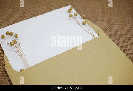 Mockup with envelope and blank card. One kraft paper envelope on burlap. Dried flowers (dry flax with seed pods) in envelope. Stock Photo