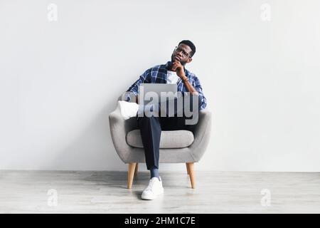 Thoughtful young black man using laptop while sitting on chair against white studio wall, full length Stock Photo