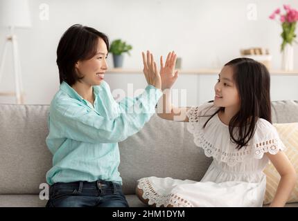 Cute little Asian girl giving high five to her grandmother, sitting on couch at home. Loving multi generation family concept Stock Photo